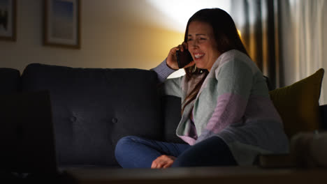 Smiling-Woman-Sitting-On-Sofa-At-Home-At-Night-Talking-On-Mobile-Phone-And-Watching-Movie-Or-Show-On-Laptop-2